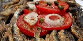 Ένα πιάτο με χρυσαφένιες, τραγανές σαρδέλες τηγανιτές, σερβιρισμένες με φέτες φρέσκιας ντομάτας.