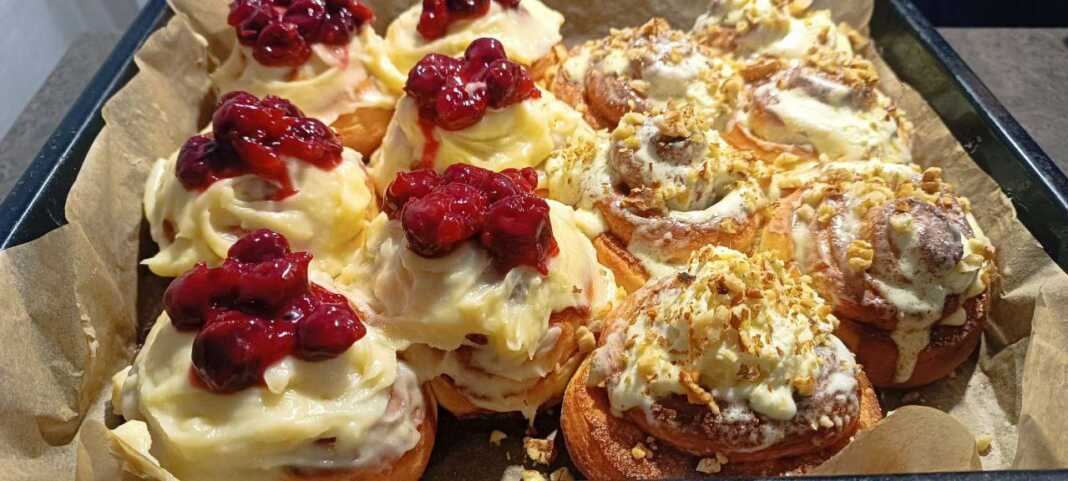 Δίσκος με αφράτα ρολά σιναμπόν καλυμμένα με λευκή κρέμα τυριού, μερικά με κανέλα και άλλα με κόκκινη γέμιση κερασιού.