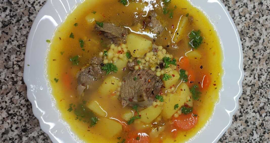 Ένα βαθύ πιάτο με αχνιστή μοσχαρόσουπα, κομμάτια τρυφερού κρέατος, πατάτες, καρότα και κουσκουσάκι, σερβιρισμένη με φρέσκο λεμόνι