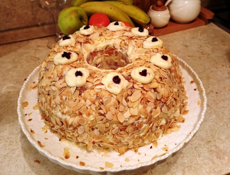 Μια εντυπωσιακή τούρτα σε σχήμα στεφανιού, καλυμμένη με λευκή κρέμα και πλούσιο καβουρδισμένο φιλέ αμυγδάλου, στολισμένη με κόκκινα κεράσια.