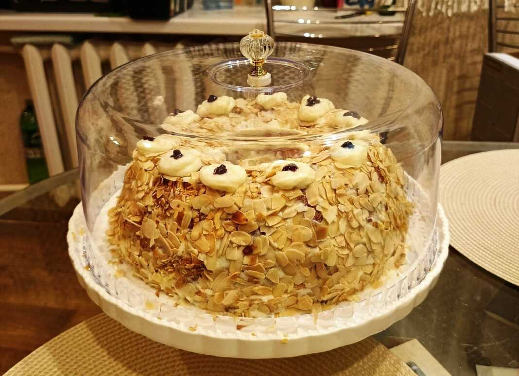 Μια εντυπωσιακή τούρτα σε σχήμα στεφανιού, καλυμμένη με λευκή κρέμα και πλούσιο καβουρδισμένο φιλέ αμυγδάλου, στολισμένη με κόκκινα κεράσια.