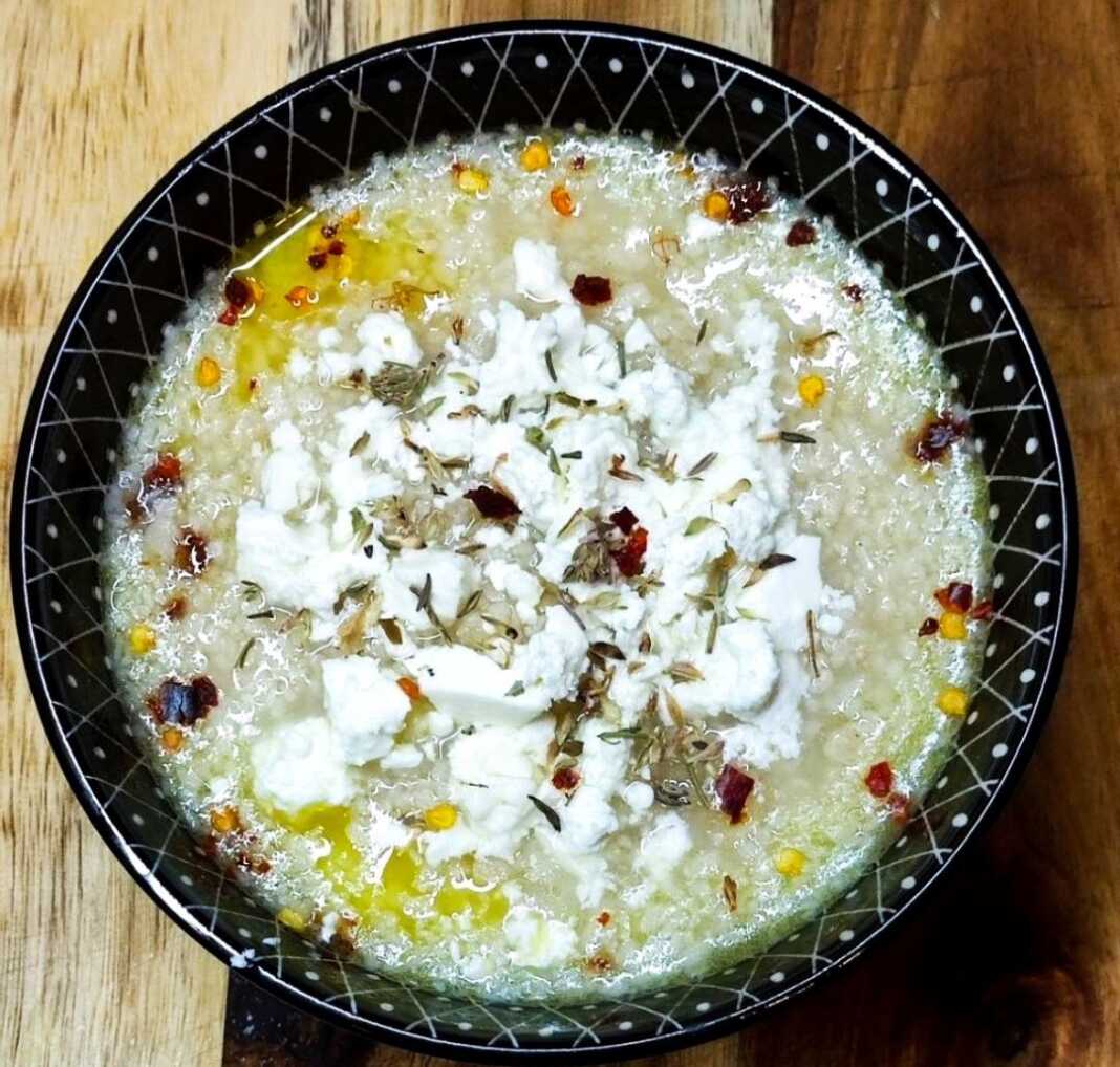 Ένα βαθύ πιάτο με αχνιστό τραχανά, πασπαλισμένο με μπόλικη τριμμένη φέτα και κόκκινο μπούκοβο, δίπλα σε μια φέτα χωριάτικο ψωμί.