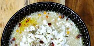 Ένα βαθύ πιάτο με αχνιστό τραχανά, πασπαλισμένο με μπόλικη τριμμένη φέτα και κόκκινο μπούκοβο, δίπλα σε μια φέτα χωριάτικο ψωμί.