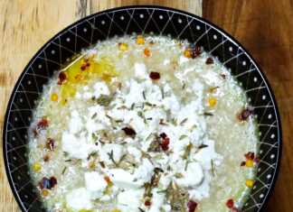 Ένα βαθύ πιάτο με αχνιστό τραχανά, πασπαλισμένο με μπόλικη τριμμένη φέτα και κόκκινο μπούκοβο, δίπλα σε μια φέτα χωριάτικο ψωμί.