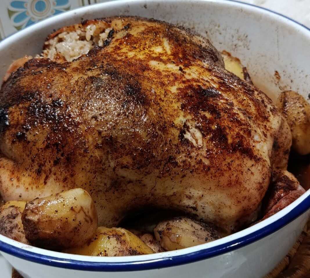 Ένα ολόκληρο ροδοκοκκινισμένο κοτόπουλο γεμιστό με ρύζι, περιτριγυρισμένο από μελωμένες πατάτες baby μέσα σε ένα κεραμικό πλακερό.