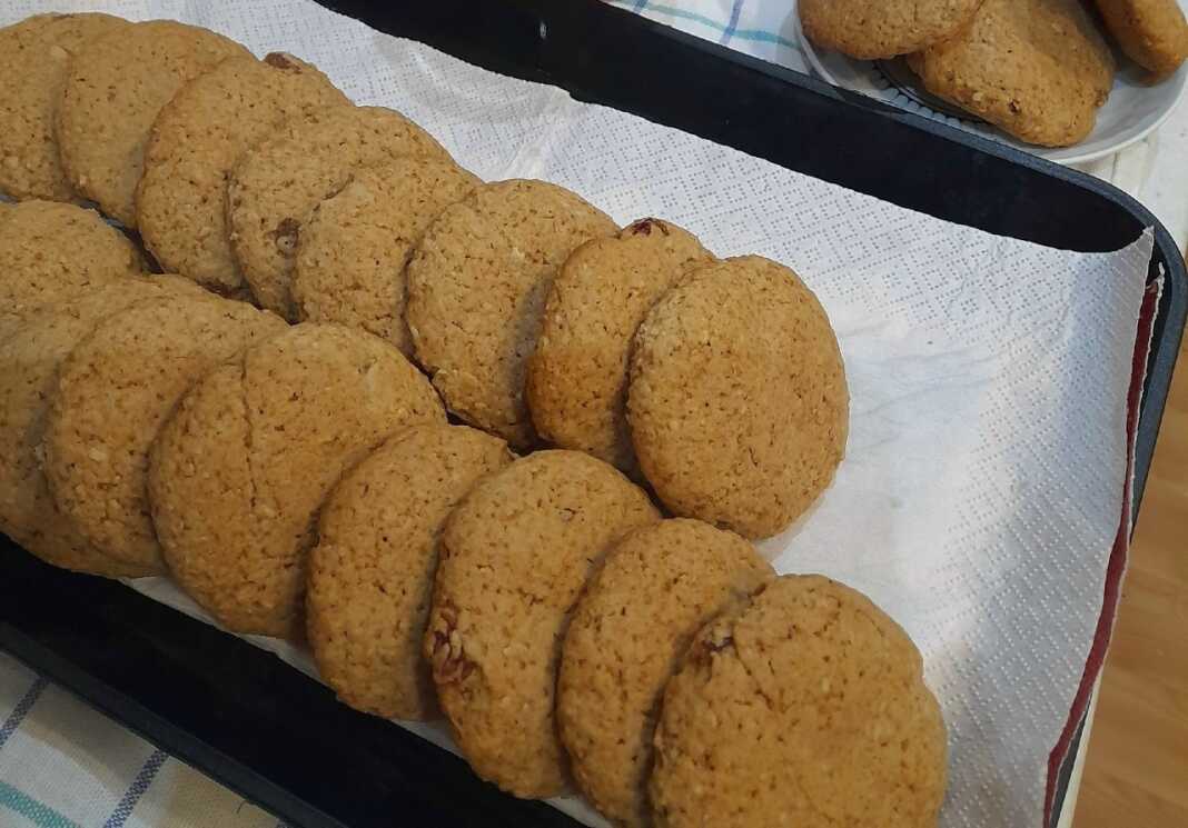 Μια πιατέλα με χρυσαφένια σπιτικά μπισκότα βρώμης, πασπαλισμένα με λίγη κανέλα και δίπλα μερικές νιφάδες βρώμης και σταφίδες.