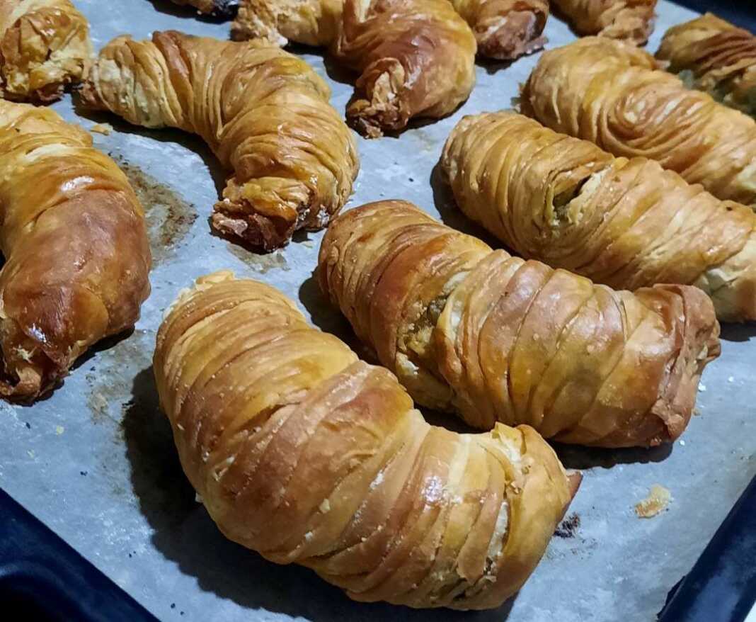 Σειρές από ροδοκοκκινισμένα σπανακοτυροπιτάκια στριφτά μέσα στο ταψί, αλειμμένα με αυγό για λάμψη