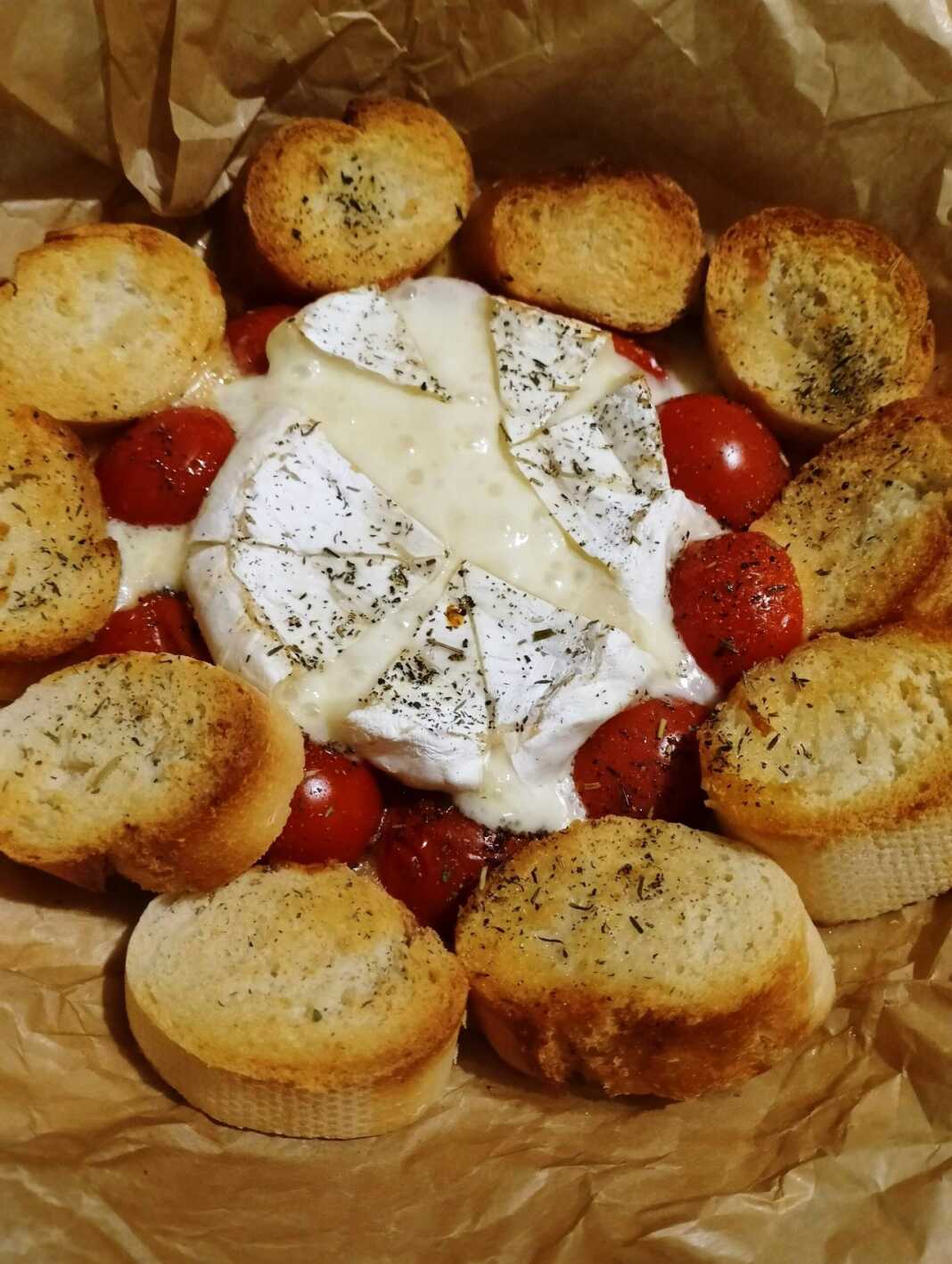 Ένα στρογγυλό ταψάκι με λιωμένο τυρί Brie στο κέντρο, περιτριγυρισμένο από ψητά ντοματίνια και τραγανές φέτες μπαγκέτας.