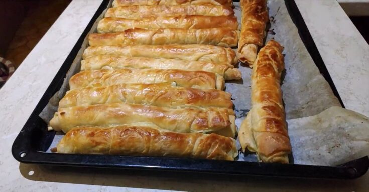 Σειρές από ροδοκοκκινισμένες χειροποίητες τυρόπιτες με σφολιάτα πάνω σε ταψί με λαδόκολλα.