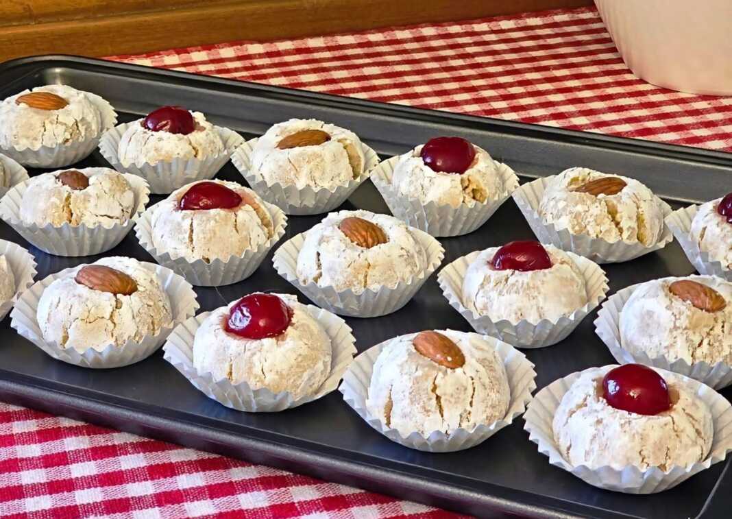 Σειρές από λευκά αμυγδαλωτά πασπαλισμένα με άχνη ζάχαρη, διακοσμημένα με ολόκληρα αμύγδαλα και κόκκινα κεράσια πάνω σε λαδόκολλα.