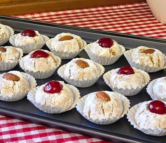 Σειρές από λευκά αμυγδαλωτά πασπαλισμένα με άχνη ζάχαρη, διακοσμημένα με ολόκληρα αμύγδαλα και κόκκινα κεράσια πάνω σε λαδόκολλα.