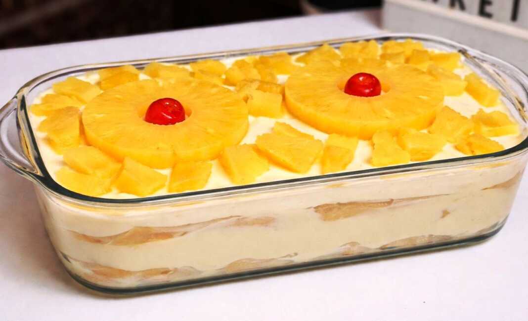 Ένα δροσερό γλυκό ψυγείου σε πυρέξ με στρώσεις σαβουαγιάρ, λευκή κρέμα και στολισμένο με ροδέλες ανανά και κόκκινα κερασάκια.