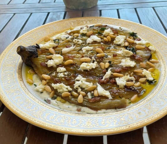 Μια ρηχή πιατέλα με μια μελωμένη ψητή μελιτζάνα τοποθετημένη πάνω σε μια λευκή κρέμα γιαουρτιού, γαρνιρισμένη με λιωμένο τυρί, κουκουνάρι και φρέσκια ρίγανη